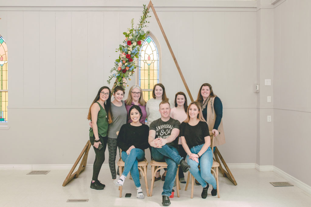 Prince Edward County photographer Deanna Way sitting in front of a triangle wedding arch with seven other individuals