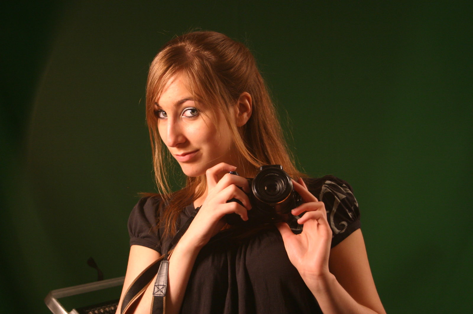 A photo of Deanna holding a camera in front of a green backdrop taken from her time in photography class