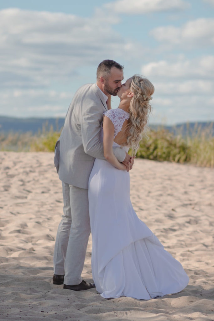 Groom in a tan suit kisses his bride on the beach after their Prince Edward County wedding reception