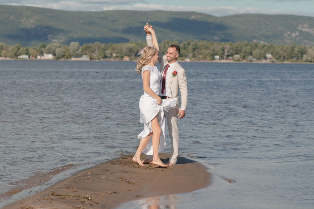 Groom in a tan suit and red tie twirls his stunning blonde bride on a sand bar next to a lake in Prince Edward County