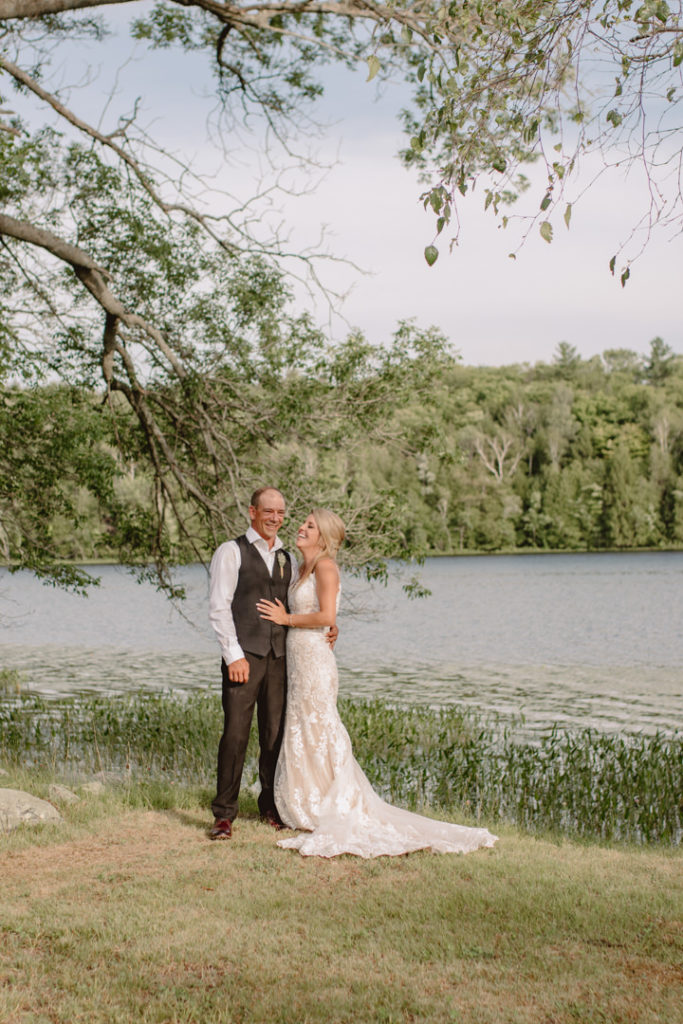 Bride and groom stand on a grassy patch under a tree in front of a lake in Prince Edward County