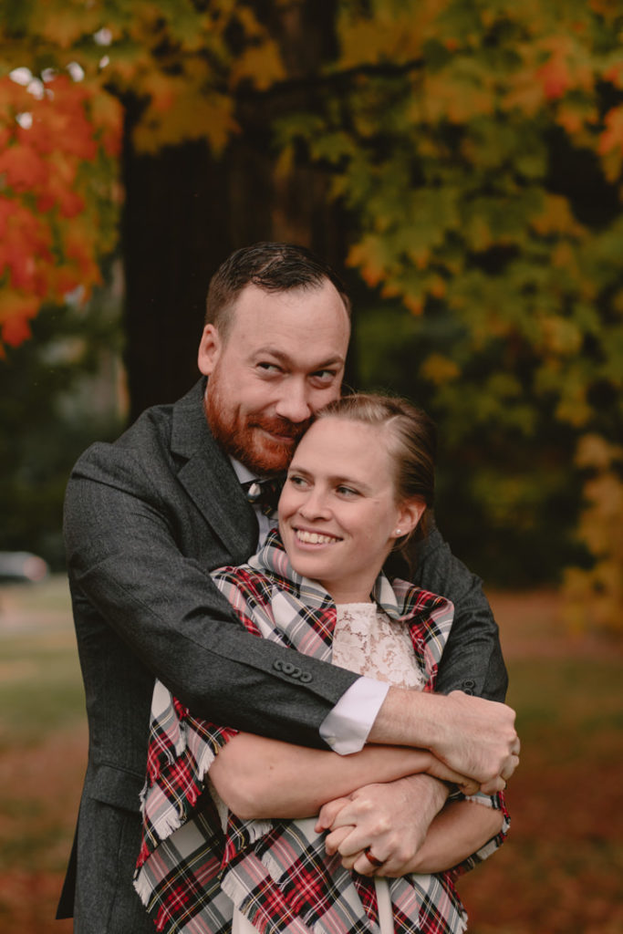 Bride in white dress and plaid shawl is held from behind by her groom in a dark grey suit during their fall wedding