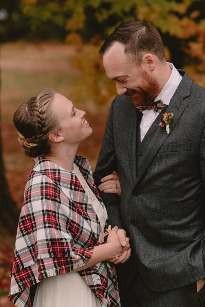 Bride in white dress and plaid shawl looks into her groom’s eyes, exchanging smiles on their fall wedding day in Ontario