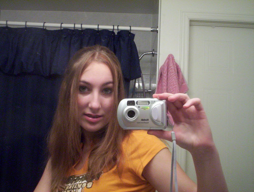 A young Deanna Way wearing a yellow t-shirt takes a mirror selfie in her bathroom with a digital camera