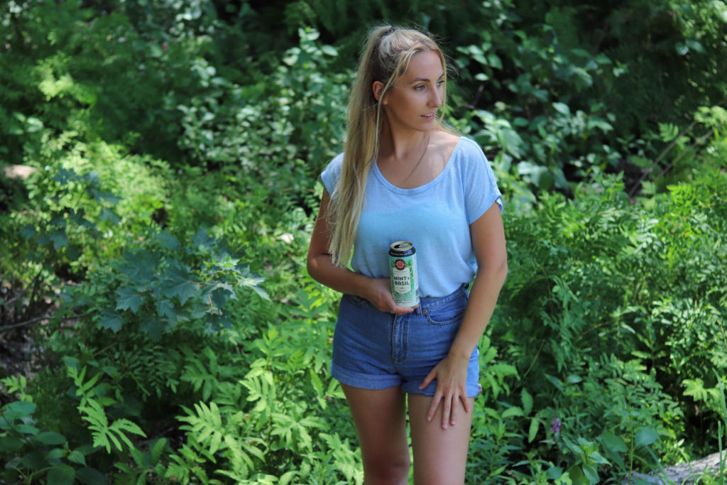 Deanna way stands in front of shrubs and greenery in jean shorts and a blue top holding a cider up in front of her