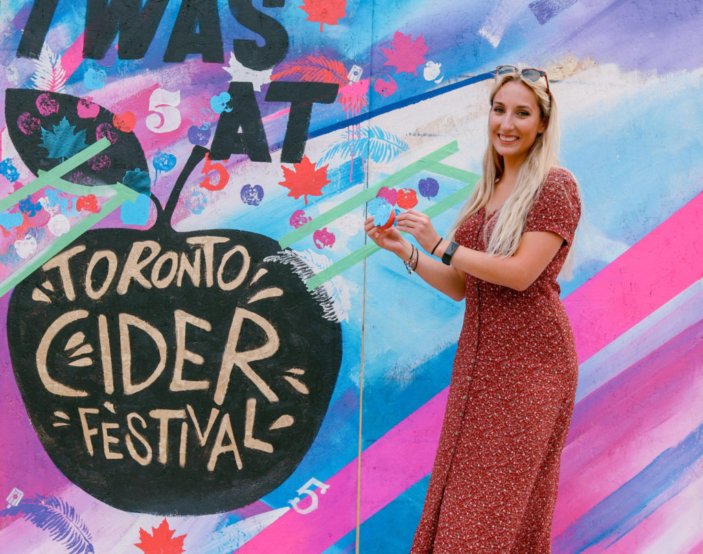 Deanna smiles in a long, button down red dress smiling in front of a Toronto Cider Festival sign in her days before becoming a professional photographer